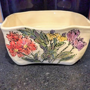 Könnte beinhalten: Eine quadratische Keramikschale mit cremefarbenem Äußeren. Die Schale ist mit einem Blumendesign mit roten, gelben und lila Blumen mit grünen Blättern verziert. Die Schale steht auf einer dunkelgrauen Oberfläche.