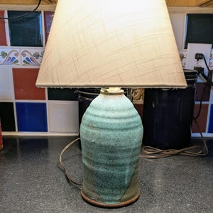 May include: A table lamp featuring a textured, cylindrical ceramic base with a mottled teal and green glaze. The lamp is topped with a tapered, off-white fabric lampshade that has a subtle cross-hatch pattern and a visible light bulb inside.