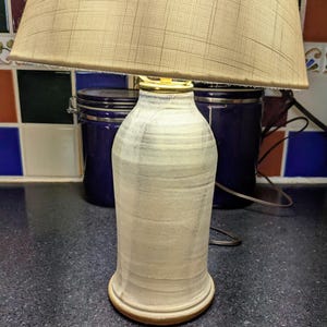May include: A table lamp featuring a cylindrical, off-white ceramic base with subtle horizontal texture and a brass-colored neck. The lamp is topped with a wide, beige fabric lampshade that has a faint, grid-like pattern woven into the material.