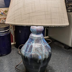May include: A table lamp featuring a hand-thrown ceramic base with a reactive glaze in shades of deep blue, black, and hints of purple. The base has a tapered neck and a wide body, topped with a textured, light beige fabric drum lampshade.