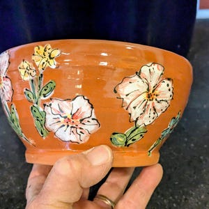 May include: Hand-painted ceramic bowl in a warm orange hue, adorned with floral designs in white, pink, yellow, and green. The bowl's exterior features detailed flower and leaf motifs, creating a rustic and artistic aesthetic. A unique piece for serving or display.
