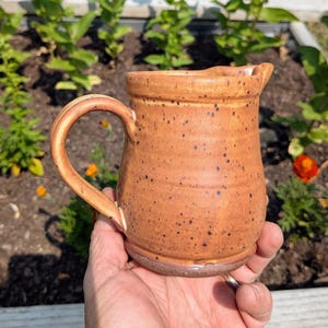 May include: A ceramic pitcher with a warm orange glaze and dark brown speckles throughout. The vessel features a rounded body, a curved handle on the side, and a small pouring spout at the top rim. The surface has a subtle textured appearance.