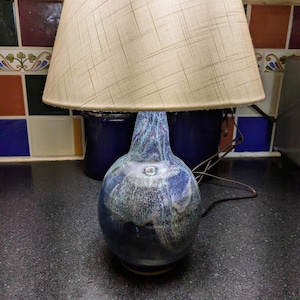 May include: A table lamp featuring a rounded ceramic base with a glossy, mottled blue and white glaze. The lamp is topped with a conical, textured beige fabric lampshade that has a subtle cross-hatched pattern across its entire surface.