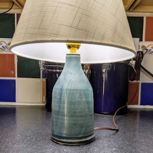 May include: A table lamp featuring a blue ceramic base with a smooth, tapered bottle-like shape and a textured, light beige fabric lampshade. The lamp includes a gold-coloured metal socket and a brown electrical cord extending from the base.