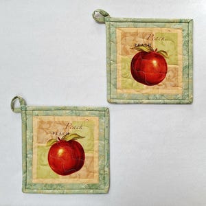 May include: Two square quilted pot holders with a red peach design. Each pot holder has a light green border and a loop for hanging. The word "Peach" is printed above the peach image.