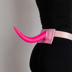 Puede incluir: Un accesorio de cola de color rosa brillante, curvado y con forma de cuerno, unido a un cinturón de color rosa claro. La cola presenta una textura lisa y sólida, montada sobre una base rectangular rosa en la parte baja de la espalda, sujeta por una correa.