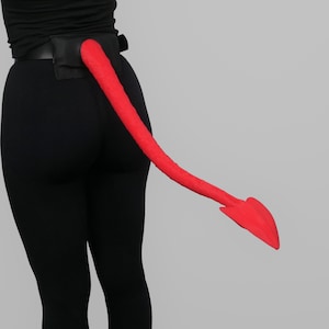 Könnte beinhalten: Ein leuchtend rotes, langes und flexibles Schwanz-Accessoire, das an einem schwarzen Gürtel befestigt ist. Der Schwanz hat eine spitze, pfeilförmige Spitze am Ende. Der Gürtel wird über einer schwarzen Leggings und einem schwarzen Oberteil getragen.