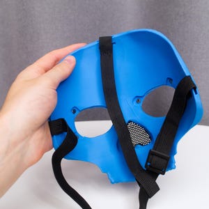 May include: A bright blue skull mask with black elastic straps and a black mesh eye covering. The mask is held by a hand, showcasing the interior and the strap design. The mask is made of a hard plastic material.