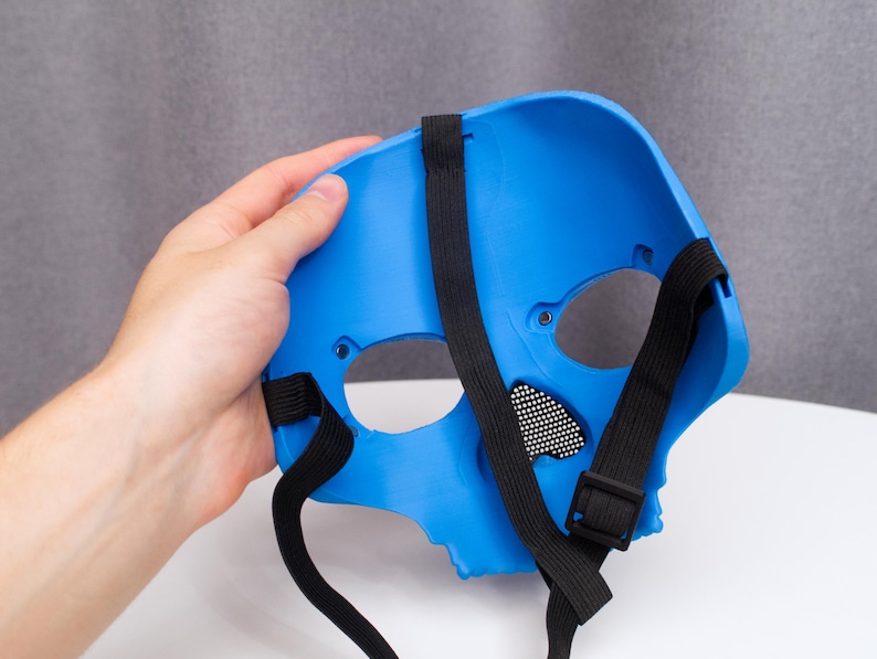 May include: A bright blue skull mask with black elastic straps and a black mesh eye covering. The mask is held by a hand, showcasing the interior and the strap design. The mask is made of a hard plastic material.