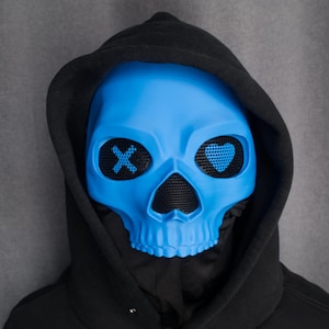 May include: A bright blue skull mask with mesh eye coverings, featuring an 'X' and a heart design. The mask is worn with a black hooded sweatshirt. The mask is a unique accessory for costumes or themed events.