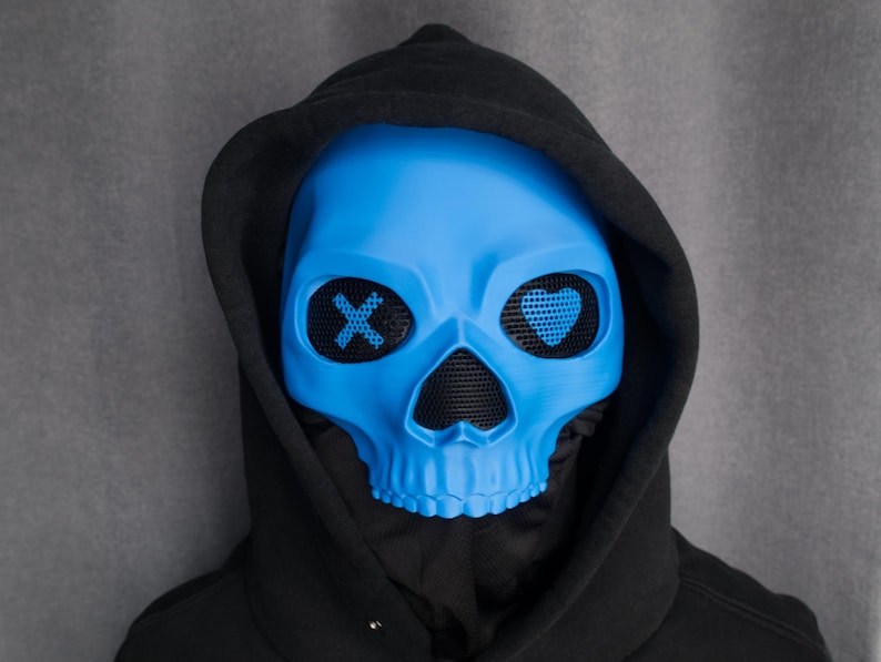 May include: A bright blue skull mask with mesh eye coverings, featuring an 'X' and a heart design. The mask is worn with a black hooded sweatshirt. The mask is a unique accessory for costumes or themed events.