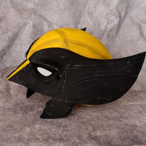 May include: A black and yellow plastic mask with a textured surface. The mask has a single eye opening and a pointed chin.