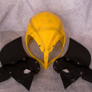 May include: A yellow helmet with a textured surface and black wings. The helmet has a pointed top and a curved front.