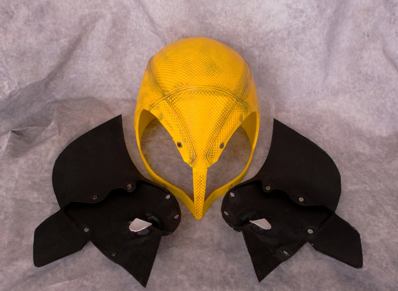 May include: A yellow helmet with a textured surface and black wings. The helmet has a pointed top and a curved front.