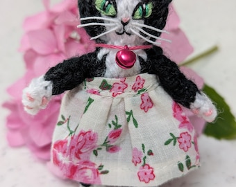 Schwarzweißes Kätzchen mit grünen Augen und schwarzem Schwanz, bekleidet mit einem cremefarbenen Rock mit rosa Rosenmuster und einem rosa Glöckchen.