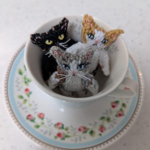 May include: Three small crocheted cat figurines in a teacup. The cats are white with gray, black with yellow, and tan with white. The teacup is white with a floral pattern.