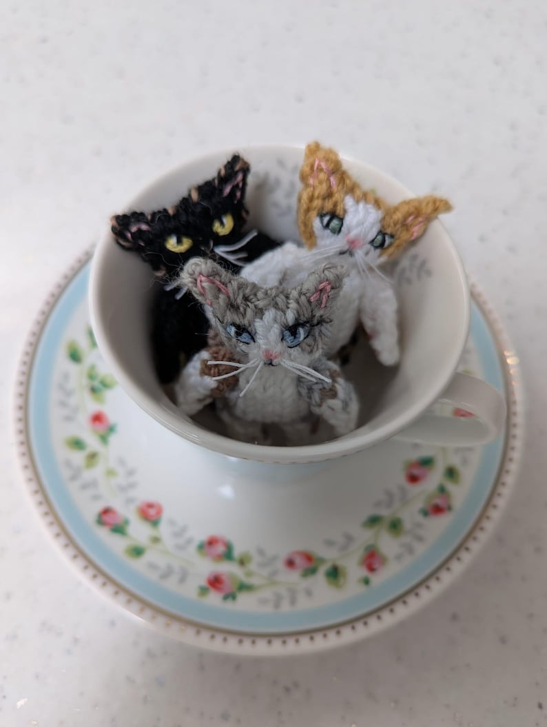 K&ouml;nnte beinhalten: Drei kleine geh&auml;kelte Katzenfiguren in einer Teetasse. Die Katzen sind wei&szlig; mit grau, schwarz mit gelb und braun mit wei&szlig;. Die Teetasse ist wei&szlig; mit einem Blumenmuster.