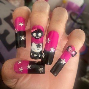 Puede incluir: Uñas acrílicas largas de color rosa y negro con un diseño de calavera, Hello Kitty y un lazo rosa. Cada uña tiene detalles de estrellas plateadas.