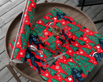 Puede incluir: Papel de regalo navideño rojo con un estampado de árboles de Navidad verdes, copos de nieve y un Bigfoot azul con gorro de Papá Noel. Un regalo envuelto con un lazo dorado y tijeras están sobre el papel.