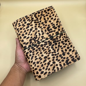 May include: A leopard print journal cover with a green elastic closure. The cover features a tan background with black spots. The journal is held in a hand.