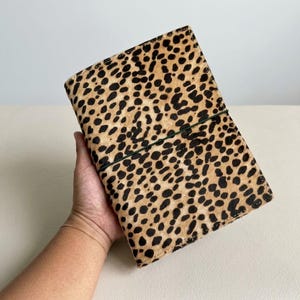 May include: A rectangular notebook cover featuring a tan and black leopard print pattern with a textured, fur-like appearance. A thin, dark green elastic band is wrapped horizontally around the centre to secure the cover closed.
