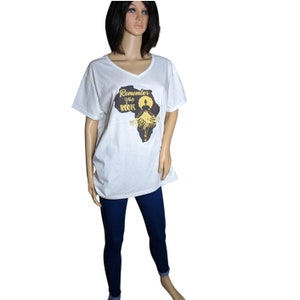 以下が含まれることがあります： Vネックの白いTシャツ。アフリカ大陸の黒いシルエットが描かれ、金色の文字で「Remember your ROOTS」と記されています。アフロヘアの人物のグラフィックが金色で施され、その髪の根元が大陸の下部に向かって伸びているデザインです。