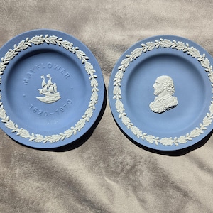 May include: Two light blue decorative plates with white relief details. One plate features a ship with the text MAYFLOWER 1620-1970. The other plate displays a white relief bust of a bearded person. Both plates have a white oak leaf border.