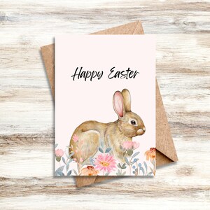 May include: A white card with the text "Happy Easter" in black lettering. A watercolor illustration of a brown rabbit with pink ears surrounded by pink and blue flowers.