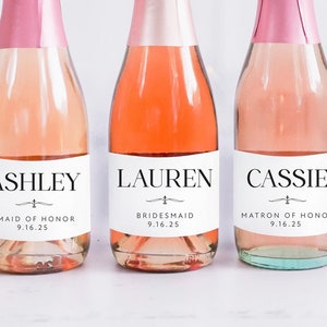 May include: Three mini bottles of sparkling wine with personalized labels. The labels are white with black text and a simple design. The bottles are labeled "Ashley Maid of Honor 9.16.25", "Lauren Bridesmaid 9.16.25", and "Cassie Matron of Honor 9.16.25".