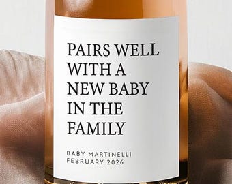 May include: A rose-colored wine bottle with a gold foil top and a white label. The label reads "PAIRS WELL WITH A NEW BABY IN THE FAMILY" with the name "BABY MARTINELLI" and the date "FEBRUARY 2026" below. The bottle sits on a pink fabric.