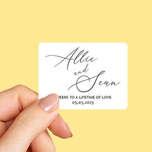 K&ouml;nnte beinhalten: Wei&szlig;er Hochzeitstags-Geschenkanh&auml;nger mit schwarzem Kalligraphie-Text, der "Allie und Sean" und "Cheers to a lifetime of love 05.03.2025" lautet.