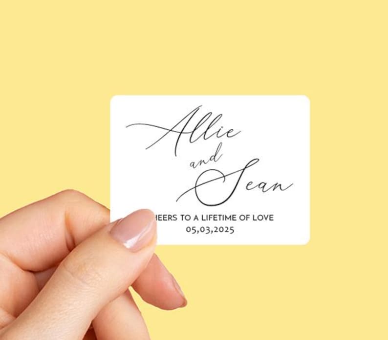 K&ouml;nnte beinhalten: Wei&szlig;er Hochzeitstags-Geschenkanh&auml;nger mit schwarzem Kalligraphie-Text, der "Allie und Sean" und "Cheers to a lifetime of love 05.03.2025" lautet.