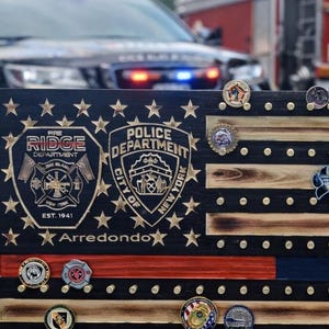 May include: Wooden American flag display with a thin blue line and a red line, holding various challenge coins. The display features the Ridge Fire Department and Police Department logos, with the text "Arredondo". A police car and fire truck are in the background.