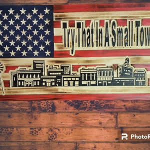 Könnte beinhalten: Eine amerikanische Holzflagge in den Farben Rot, Weiß und Blau. Die Flagge hat eine Silhouette einer Stadtlandschaft mit dem Text "Try That In A Small Town" in schwarzen Buchstaben.