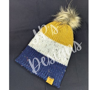 Puede incluir: Un gorro de punto a rayas con un pompón de piel sintética. El gorro es azul, blanco y amarillo.