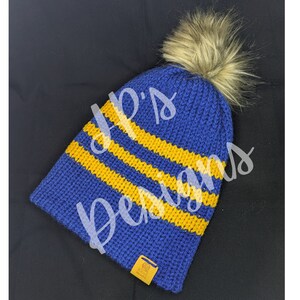 May include: A blue knit beanie with three yellow stripes and a faux fur pom-pom. The hat has a small tag with text. The text "P.S. Designs" is written across the hat in a cursive font.