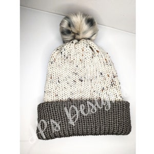 May include: A white knit beanie with a brown cuff and a fluffy white pom pom on top. The beanie has a speckled pattern throughout.