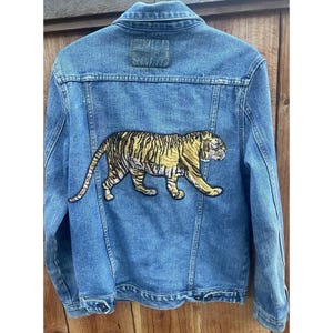 May include: A blue denim jacket featuring a large embroidered patch of a tiger on the back. The tiger is depicted in profile, walking toward the right, with yellow and black stripes. The jacket has a classic collar, long sleeves, and buttoned cuffs.