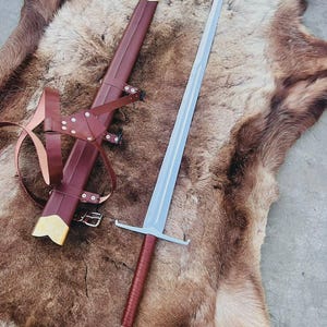 Op de afbeelding: Een langzwaard met een zilverkleurig lemmet, een met bruin leer omwikkelde greep en een metalen pommel. Daarnaast ligt een bijpassende bruin lederen schede met goudkleurige metalen beslag aan de punt en een bruin lederen harnas met zilveren klinknagels.