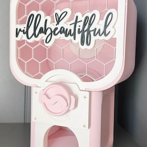 Könnte beinhalten: Ein rosa-weißer Kaugummiautomat mit einem Hintergrund im Wabenmuster. Auf der Vorderseite befindet sich der schwarze Schriftzug "villabeautiful", der auf einem weißen Hintergrund liegt. Er verfügt über einen Drehknopf und eine Ausgabeschale.