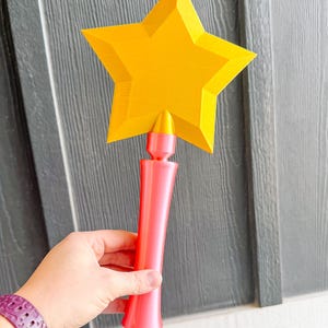 May include: A star-shaped wand with a yellow star and a pink handle. The wand is held against a grey and white background. The star is a bright yellow colour, and the handle is a vibrant pink. The wand appears to be 3D printed.