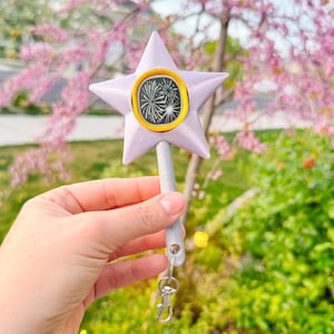 May include: A lavender star-shaped wand with a gray handle and a gold-rimmed center displaying a firework design. The wand has a silver clasp attached to the handle. The background features a blurred view of a tree with pink blossoms and green foliage.