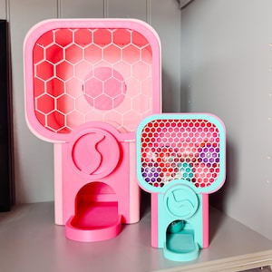 May include: Two pink and teal gumball machines with a honeycomb design on the front. The larger machine is all pink, while the smaller one has a teal frame and dispenser. Both have a curved design on the front.