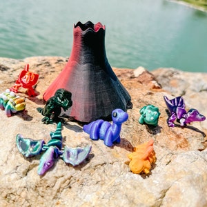 May include: A collection of colorful dinosaur figurines, including a green dinosaur, a purple dinosaur, a red dinosaur, a blue dinosaur, and a green dinosaur with a black volcano in the background.