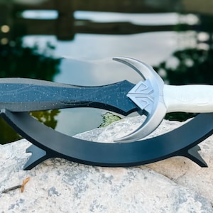 Könnte beinhalten: Ein schwarzes und silbernes Schwert mit einem Halbmond-Design am Griff, das auf einem schwarzen Ständer präsentiert wird. Das Schwert liegt auf einem grauen Stein.