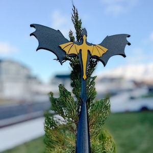 May include: A black and gold dragon garden ornament. The dragon has outstretched wings and a long, pointed tail. The ornament is inserted into a green bush, with a blurred background of a blue sky and houses.