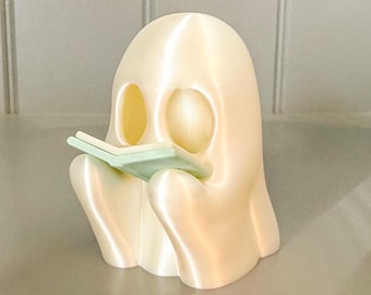 May include: A 3D-printed ghost figurine in a sitting position, holding a small, light green book. The ghost is off-white with two oval eye holes and a curved body. The book is held in its lap.