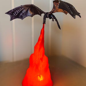 May include: A black dragon figurine with outstretched wings, flying over a red flame base. The flame base is lit from within, creating a warm glow.