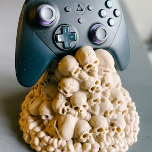 Op de afbeelding: Een zwarte videogamecontroller met paarse knoppen staat op een witte schedelvormige standaard. De standaard is gemaakt van veel kleine schedels die op elkaar gestapeld zijn.