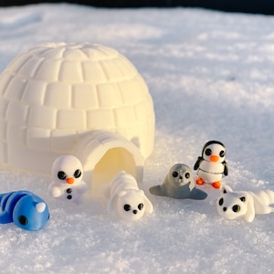 May include: A white igloo with a small penguin, a snowman, a seal, and two arctic foxes on a snowy surface.
